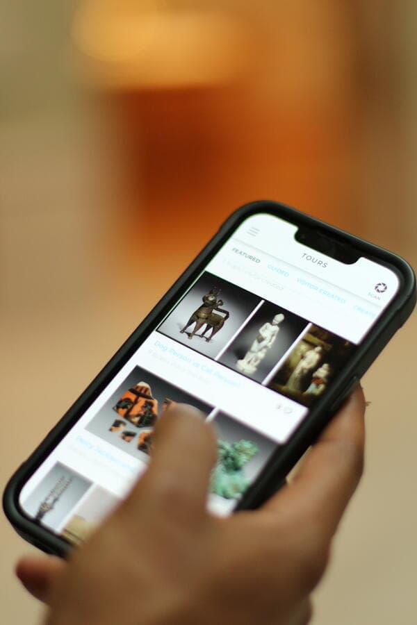 A hand holding a phone with the Artlens App, using their thumb to browse artwork images.