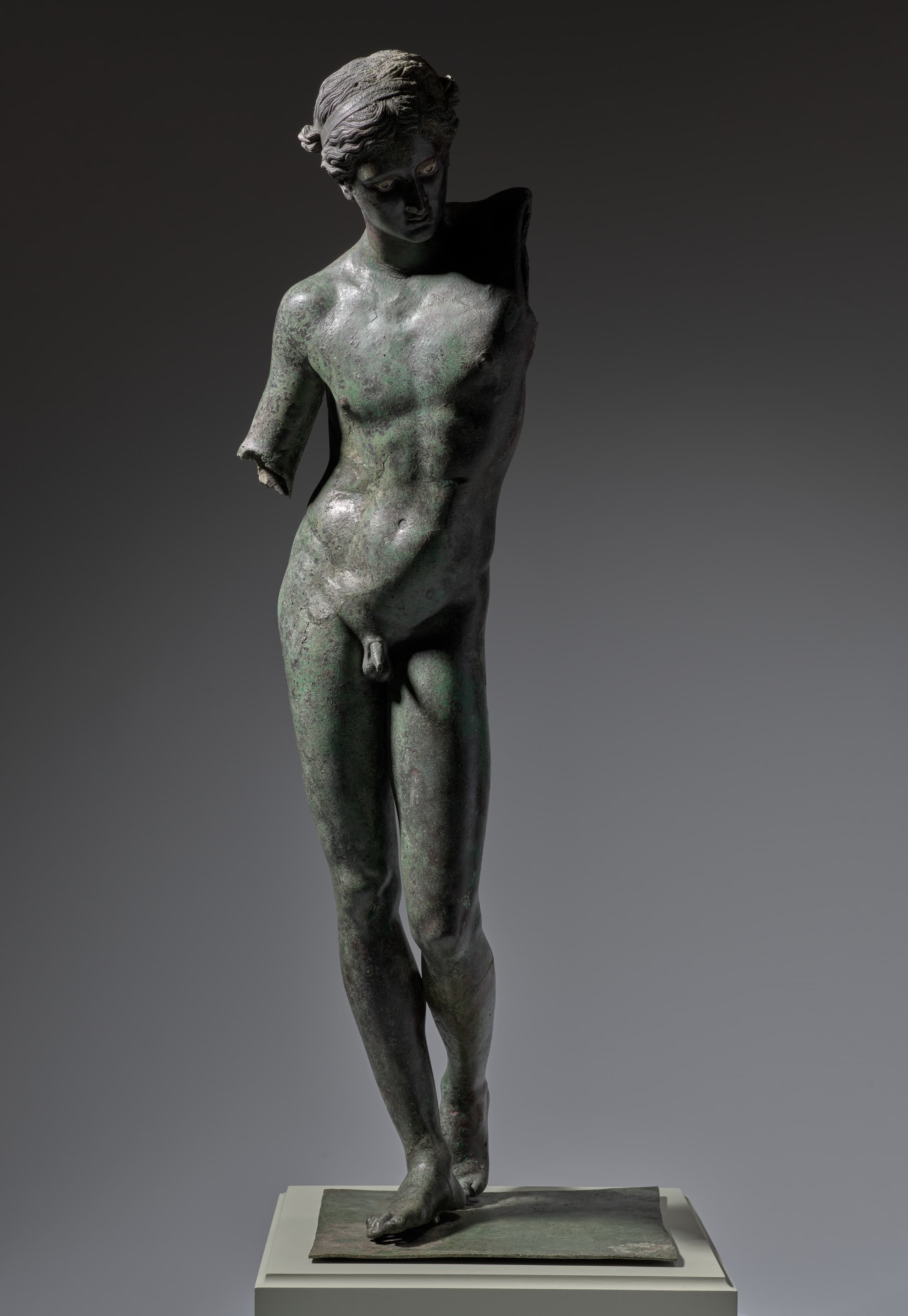 A life-size, dark gray, bronze sculpture of a nude male standing. His head is looking slightly down to our right, his genitalia is shown, his hips are cocked to the side, and his left leg is slightly bent. The arms of the sculpture are broken off, with his left arm broken off from the shoulder and his right arm broken off from his elbow.