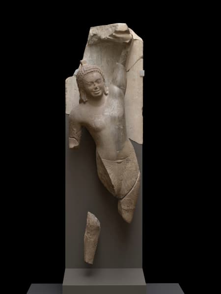 Fragmented grey sandstone sculpture of Krishna in human form, reaching his left arm up, right arm hanging down and broken away at the bicep. For the legs, only a fragment of the right shin remains, a skirt with radiating arms wrapped around the waist and bare chested. Krishna smiles faintly, face worn down, ears elongated, and hair falling to shoulders in tightly coiled curls.