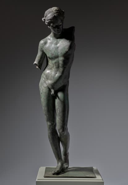 A life-size, dark gray, bronze sculpture of a nude male standing. His head is looking slightly down to our right, his genitalia is shown, his hips are cocked to the side, and his left leg is slightly bent. The arms of the sculpture are broken off, with his left arm broken off from the shoulder and his right arm broken off from his elbow.