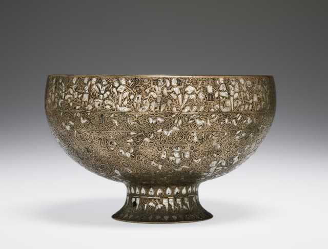 A brass, bowl-shaped cup covered in intricate designs. The rim of the cup features inscriptions of letters composed of human and animal figures. All around the cup are interlacing bands filled with human and animal figures.
