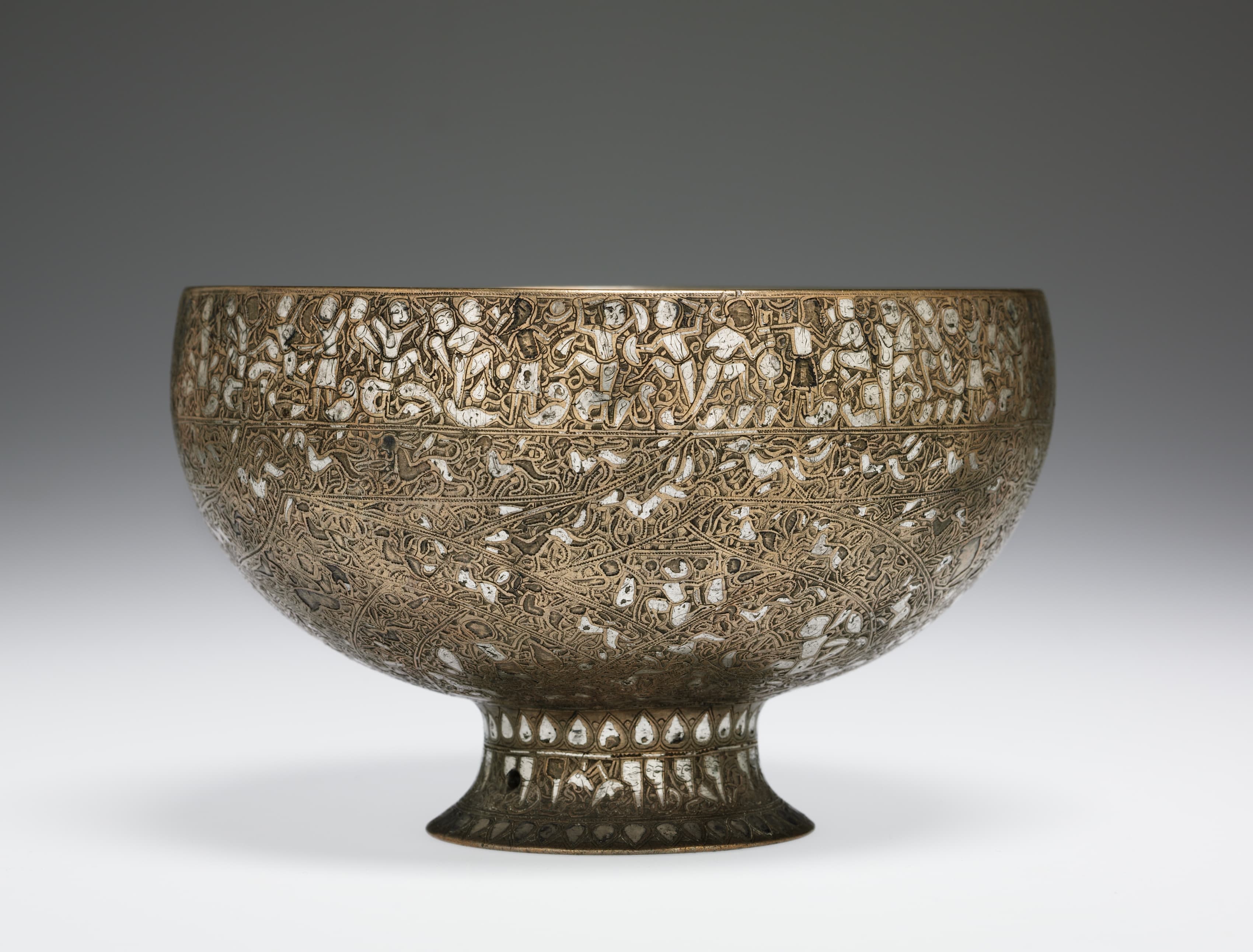 A brass, bowl-shaped cup covered in intricate designs. The rim of the cup features inscriptions of letters composed of human and animal figures. All around the cup are interlacing bands filled with human and animal figures.